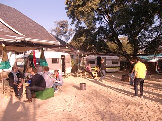 Das Bild zeigt den Parkplatz des Goethe-Instituts. Mehrere Menschen sind vor zwei Wohnwagen versammelt. Der Boden ist mit Sand bedeckt, und Bäume spenden Schatten über dem Bereich. Einige Personen sitzen auf improvisierten Sitzgelegenheiten aus Holzpaletten, während andere stehen, umhergehen oder Fußballspielen . Auf der linken Seite des Bildes ist die alte Villa mit schrägem Dach zu sehen.