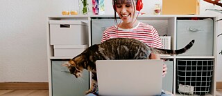 Eine junge Frau mit Laptop und rotem Kopfhörer lächelt, während eine Katze über die Tastatur geht