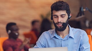 Ein Mann mit Headset sitzt hinter einem Laptop und tippt lächelnd