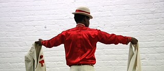 Eine Person steht mit dem Rücken zur Kamera. Sie trägt ein rotes Oberteil und einen weißen Hut mit rotem Band. In der linken Hand hält die Person ein weißes Sakko mit kariertem Muster, in der rechten Hand eine passende weiße Hose mit demselben Muster. Der Hintergrund besteht aus einer weißen Ziegelwand.