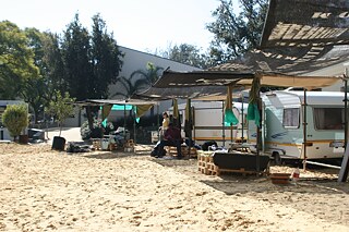 Das Bild zeigt eine sandige Fläche, auf der mehrere Wohnwagen nebeneinander geparkt sind. Vor den Wohnwagen spenden provisorische Sonnendächer aus Stoff, die mit Stangen befestigt sind, Schatten über Sitzbereiche. Unter den Überdachungen sitzen Menschen, und rundherum stehen verschiedene Gegenstände wie Stühle, Tische und Kisten. Im Hintergrund sind Bäume und ein Gebäude zu sehen.