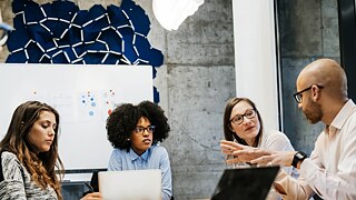 Junge Frauen und Männer sitzen zu einer Besprechung an einem Tisch. Ein Mann spricht und gestikuliert