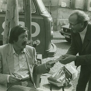 La photo en noir et blanc montre Günter Grass assis sur une chaise en pleine rue, pipe à la bouche, en pleine campagne électorale. Il tend un livre à un homme souriant penché vers lui. 