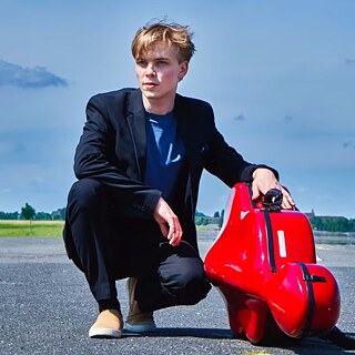 On voit Philipp Schupelius, un genou posé au sol, sur une surface asphaltée qui évoque une piste d’atterrissage ou une route dégagée. Il porte un costume noir avec une chemise foncée et des chaussures claires. À ses côtés se trouve un étui à violoncelle rouge vif. À l’arrière-plan, le ciel est vaste, parsemé de quelques nuages, avec des champs verts à l’horizon.