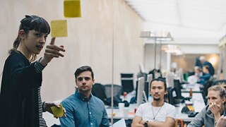 Eine junge Frau steht und präsentiert Inhalte anhand von Postits an einer Glaswand, dahinter sitzen Zuhörende