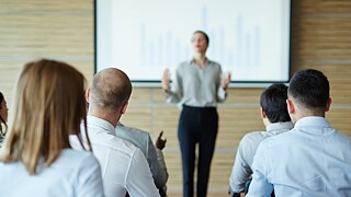 Blick von hinten in einen Seminarraum mit Publikum. Vorne steht eine Frau vor einer Leinwand und spricht