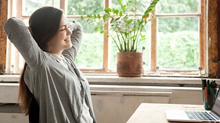 Eine junge Frau sitzt lächelnd und mit geschlossenen Augen vor einem Laptop an einem Tisch. Sie hat die Hände hinter ihrem Kopf verschränkt.