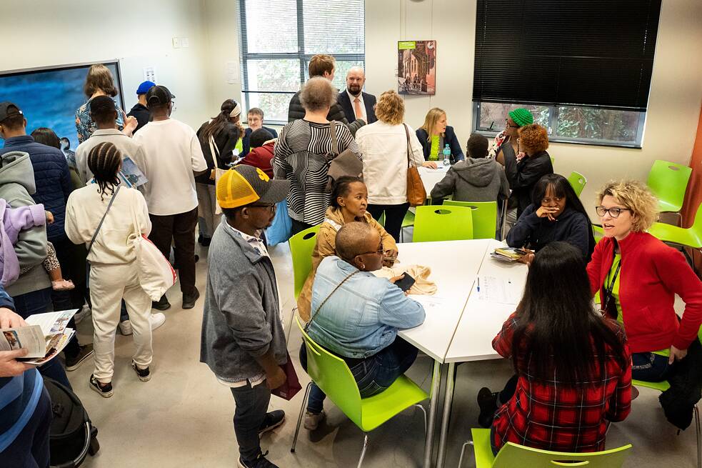 Das Bild zeigt eine Gruppe von Menschen in einem Raum mit grünen Stühlen und Tischen. Einige Personen sitzen an den Tischen, während andere stehen und sich unterhalten oder umsehen. Der Raum ist hell, mit großen Fenstern, durch die Tageslicht einfällt.