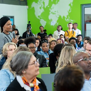 The picture shows a group of people sitting in a room. One person is standing and holding a microphone - they appear to be asking a question or making a statement. A green wall with a world map painted on it can be seen in the background. The scene looks like a lecture.