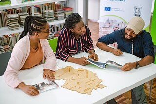 The picture shows three people sitting at a table in a library. They are looking at books and a wooden puzzle or map lying on the table. In the background are bookshelves filled with numerous books. There are some posters on the wall.