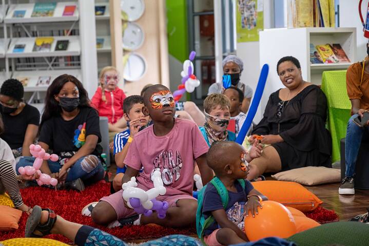 The picture shows a group of people, including children and adults, sitting on the floor in the library. Some people are holding balloon figures in their hands. In the background are bookshelves with various books and objects. Some people are sitting on cushions and the floor is covered with a red carpet.