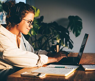 Videokonferenz im Homeoffice. Die Sprecherin gestikuliert; am Bildschirm sind weitere Konferenzteilnehmer zu sehen, sie lächeln freundlich und hören aufmerksam zu. Im Bildfokus sind die Gesprächspartner auf dem geteilten Monitor.