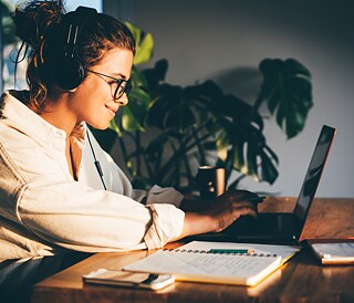 Videokonferenz im Homeoffice. Die Sprecherin gestikuliert; am Bildschirm sind weitere Konferenzteilnehmer zu sehen, sie lächeln freundlich und hören aufmerksam zu. Im Bildfokus sind die Gesprächspartner auf dem geteilten Monitor.