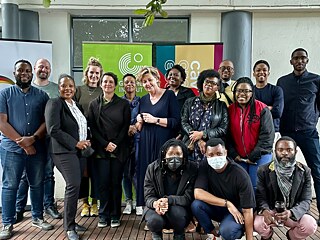 A group of people pose for a photo on a wooden platform or terrace. Two banners can be seen in the background - one bears the words “giz”, the other shows logos and text that are only partially legible. The people are arranged in two rows: Some are standing, others are kneeling or squatting. The clothing ranges from casual to formal.