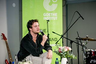 A person is sitting on a stage holding a microphone. In the background is a green banner with the words “Goethe-Institut”. Musical instruments such as a guitar and a drum kit can also be seen on the stage. A table with bottles of water and a vase of flowers stand in front of the person.