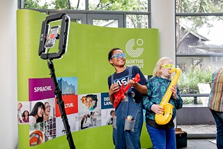 Two children stand in front of a green pull-up banner of the Goethe-Institut. Children are holding inflatable musical instruments - a red guitar and a yellow saxophone. In front of them is a ring light with a smartphone attached to it, indicating that they are being photographed or filmed.