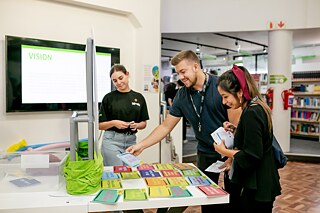 Three people are standing around a table on which numerous colorful cards are spread out. One person is pointing at the cards, another is holding papers. A screen and shelves with books can be seen in the background.