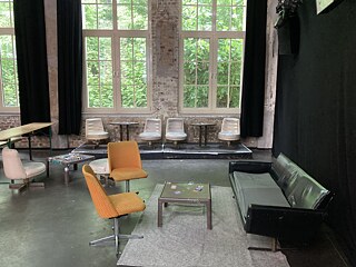 Innenraum mit großen Fenstern, schwarzem Sofa rechts. zwei orangefarbenen Stühlen links und kleinen runden Tisch dazwischen, sowie einigen weißen Stühlen im Hintergrund