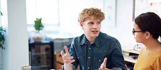 Ein junger Mann sitzt im Büro neben einer Kollegin. Die beiden unterhalten sich angeregt.