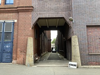 Eingang des Atomino zwischen zwei Backsteingebäuden; links befindet sich eine blaue Tür mit Graffiti, rechts ein Schild mit der Aufschrift ‚ATOMINO‘ und einem Pfeil nach links. 