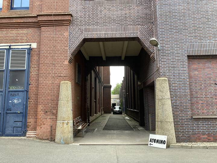 Eingang des Atomino zwischen zwei Backsteingebäuden; links befindet sich eine blaue Tür mit Graffiti, rechts ein Schild mit der Aufschrift ‚ATOMINO‘ und einem Pfeil nach links. 