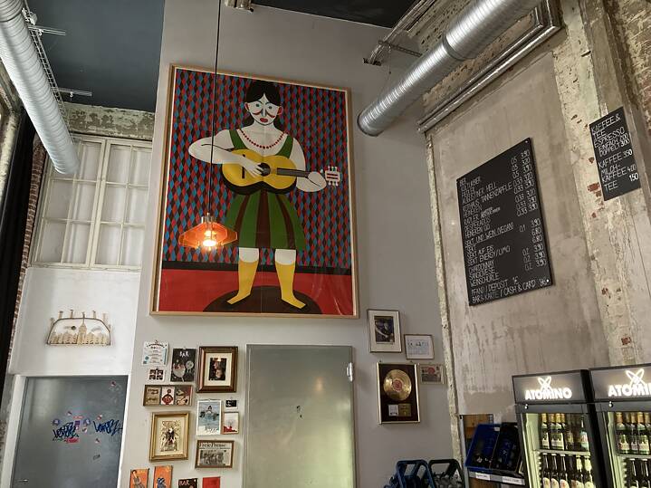 Café-Innenraum mit Wandkunst einer Gitarrenfigur, gerahmten Bildern, goldener Schallplatte, Speisekarte und zwei ‚ATOMINO‘-Kühlschränken