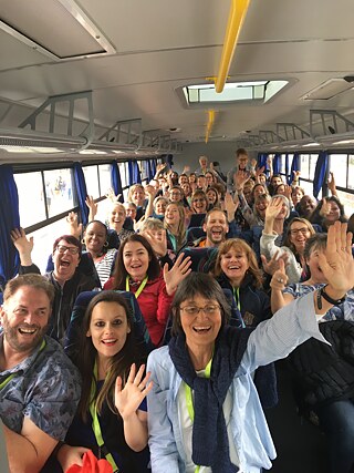 The picture shows a group of people sitting on a bus. Many of them have their hands raised and are waving. The bus is equipped with blue curtains on the windows, luggage racks above the seats and a yellow grab rail along the ceiling.
