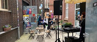 Drei skulpturale Figuren aus runden und zylindrischen Metallteilen stehen in einer Reihe auf einem gepflasterten Platz im Freien. Die mittlere Figur trägt ein rotes Oberteil und blaue Hosen, während die äußeren Figuren schwarze Beine haben