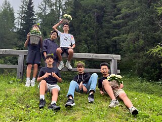 Eine kleine Gruppe Jugendlicher auf einer Alm, sie halten z.T. Pflanzen in der Hand und tragen Kränze auf dem Kopf.