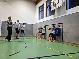 Junge Menschen lehnen in Sitzposition an einer Wand in einer Sprothalle. Die Arme sind vor dem Körper gestreckt.