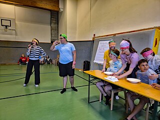 Jugendliche sitzen an einer Bierbank, sie tragende schützende Plastikfolien. Andere stehen hinter ihnen mit verbundenen Augen.