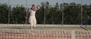 Schauspieler Sebastian Schneider im weißen Tenniskostüm mit Sonnenbrille und Schläger in der Hand