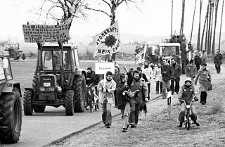 Schwarz-weiß Bild: Menschen in Traktoren, zu Fuß und auf Fahrrädern protestieren mit Schildern auf denen steht "Atomkraft, nein danke"