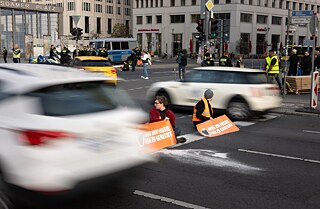 Zwei Menschen sitzen in der Mitte einer befahrenen Straße, Sie haben orange Westen an  und orange Schilder.
