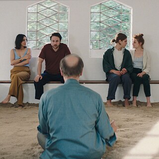 2 couples are sitting  on a bench in a white room, a man with his back to us sits facing them.