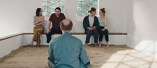 2 couples are sitting  on a bench in a white room, a man with his back to us sits facing them.