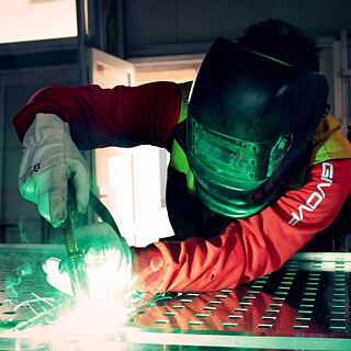 Person wearing a welding helmet and gloves, focused on welding a metal surface.