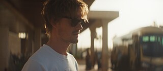 Im Fokus auf der linken Bildseite: der Protagonist Tom mit Sonnenbrille und weißem T-Shirt, nachdenklich vor dem Eingang der Hotelanlage. Rechts verschwommen im Hintergrund: ein Bus, aus dem gerade neue Besucher aussteigen.