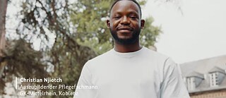 Christian Njiotch, a nursing trainee, standing outdoors with trees and a building in the background.