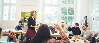 Klassenraum mit Studenten. Die Lehrerin steht in der Mitte und lächelt. Die Studenten heben die Hände.