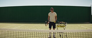 Tom auf dem Tennisplatz mit einem einem Wagen voller Tennisbälle.