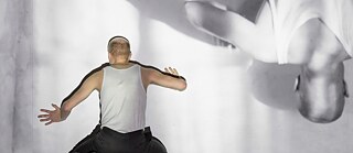 Tänzer steht vor Wand mit dem Rücken zum Publikum. Sein Bild wird auf dem Kopf stehend auf dieselbe Wand projiziert.