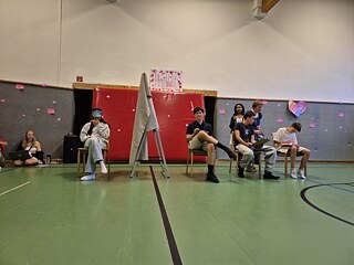 Jugendliche sitzen auf Stühlen in einer Sporthalle, es trennt Sie eine Wand aus Stoff. Einer hat die Augen verbunden.