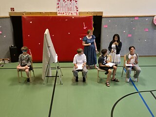 Jugendliche sitzen auf Stühlen in einer Sporthalle, es trennt Sie eine Wand aus Stoff. Einer hat die Augen verbunden.