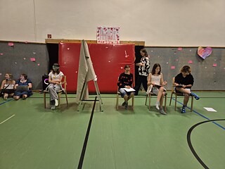 Jugendliche sitzen auf Stühlen in einer Sporthalle, es trennt Sie eine Wand aus Stoff. Einer hat die Augen verbunden.