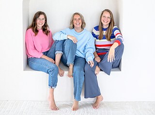 Drei weiße Frauen Anfang Dreißig sitzen in einer weißen Sitzecke. Sie lächeln in die Kamera.