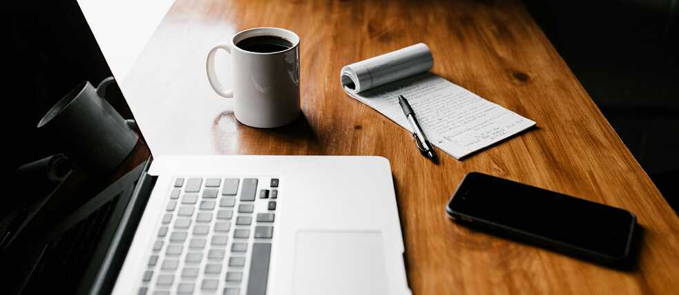 A wooden desk with a laptop, a cup of coffee, a notepad with handwritten notes, a black pen, and a smartphone. The workspace is well-lit, creating a cozy and focused atmosphere.