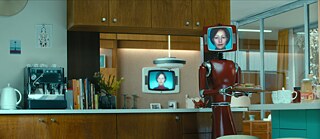 Against the backdrop of a 1970s kitchen unit, Cassandra - a dark red robot with an attached tube screen showing a white woman with neck-length hair - holds a plate of sandwiches. 