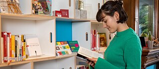 Eine junge Frau in einem grünen Pullover steht vor einem Bücherregal und hält ein aufgeschlagenes Buch, in dem sie liest, in den Händen.