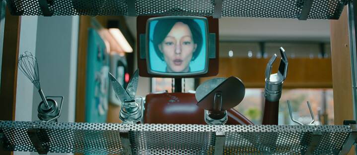 Cassandra - a dark red robot with an attached tube screen showing a white woman with neck-length hair views sharp kitchen utensils that attach to her arms.